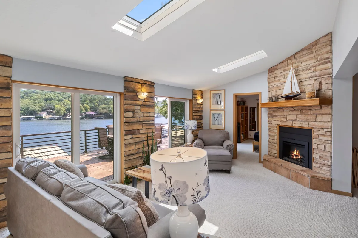 A bright, open living room featuring a stunning stone fireplace, expansive windows offering panoramic lake views, and a comfortable seating area with a decorative lamp. Natural light fills the space, highlighting the tranquil and welcoming atmosphere.