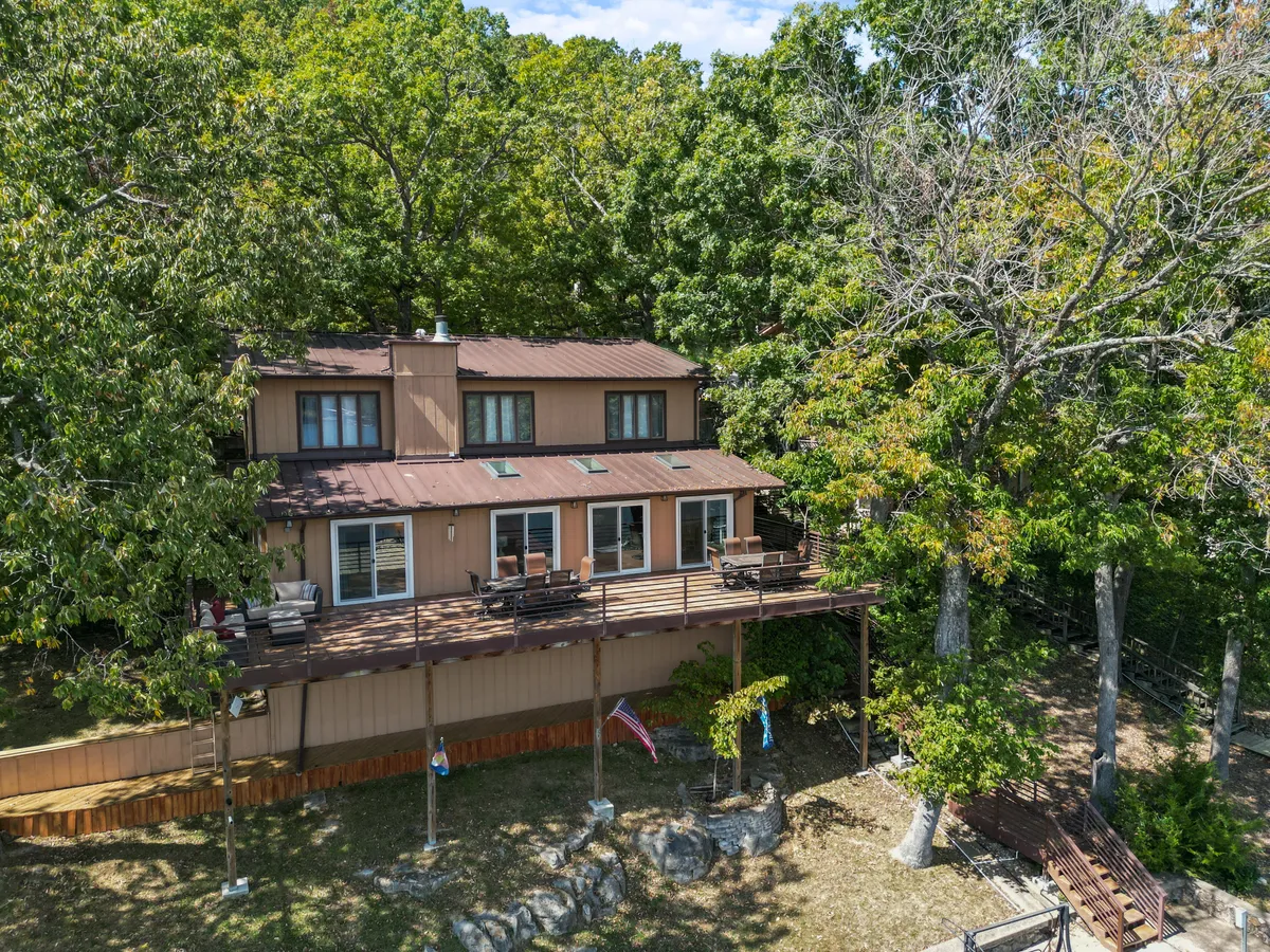 A welcoming wooden deck surrounded by large windows and lush trees, adorned with flag decorations. The area is set up for outdoor gatherings, with ample seating and a tranquil setting perfect for relaxing, socializing, or enjoying a barbecue with family and friends.
