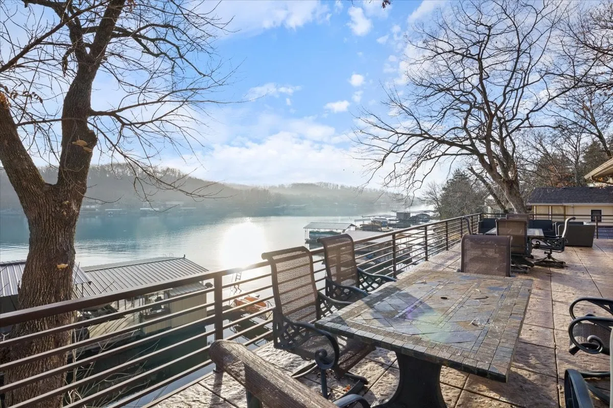A tranquil lakeside setting featuring comfortable outdoor seating and a table overlooking the water. Trees frame the scene, and distant hills create a peaceful backdrop, inviting guests to relax, dine al fresco, or socialize while enjoying beautiful lake views.