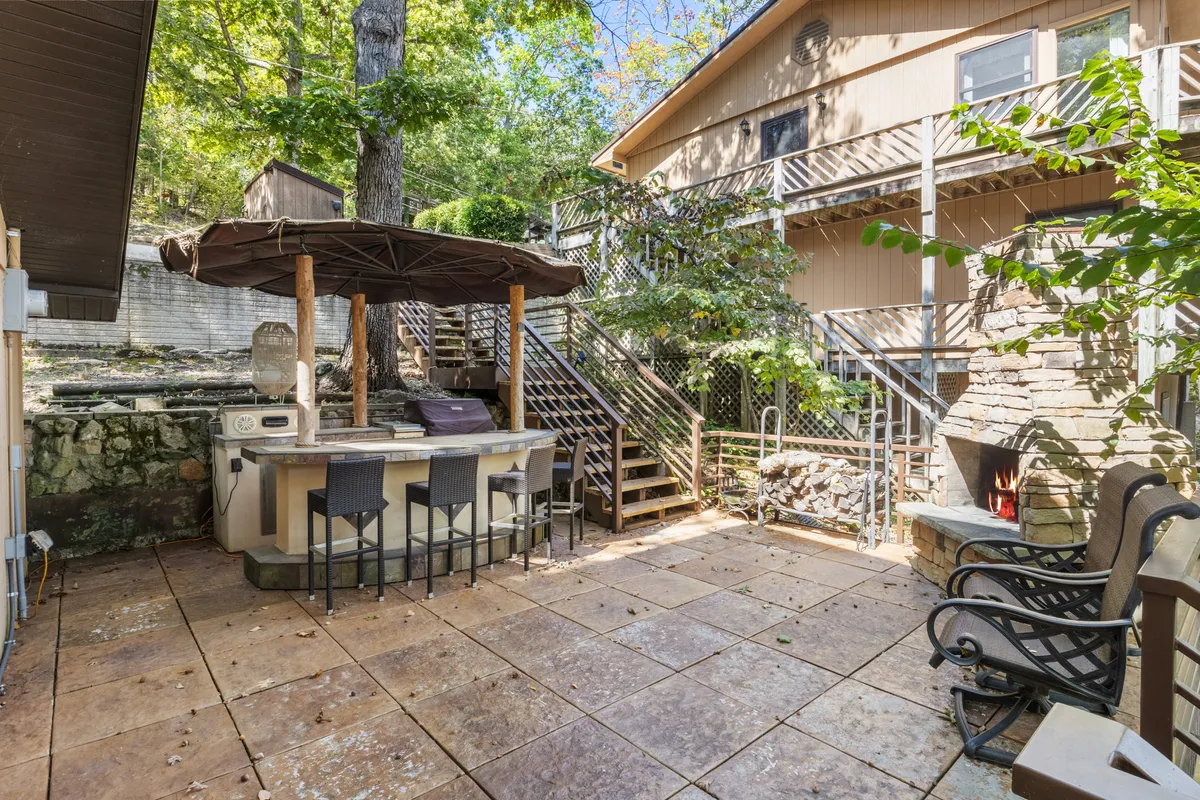 An inviting outdoor bar area nestled beside natural stone features, with ample seating and a staircase leading down to more space. Warm lighting enhances the festive mood, and the setup is ideal for hosting gatherings or enjoying drinks at sunset.