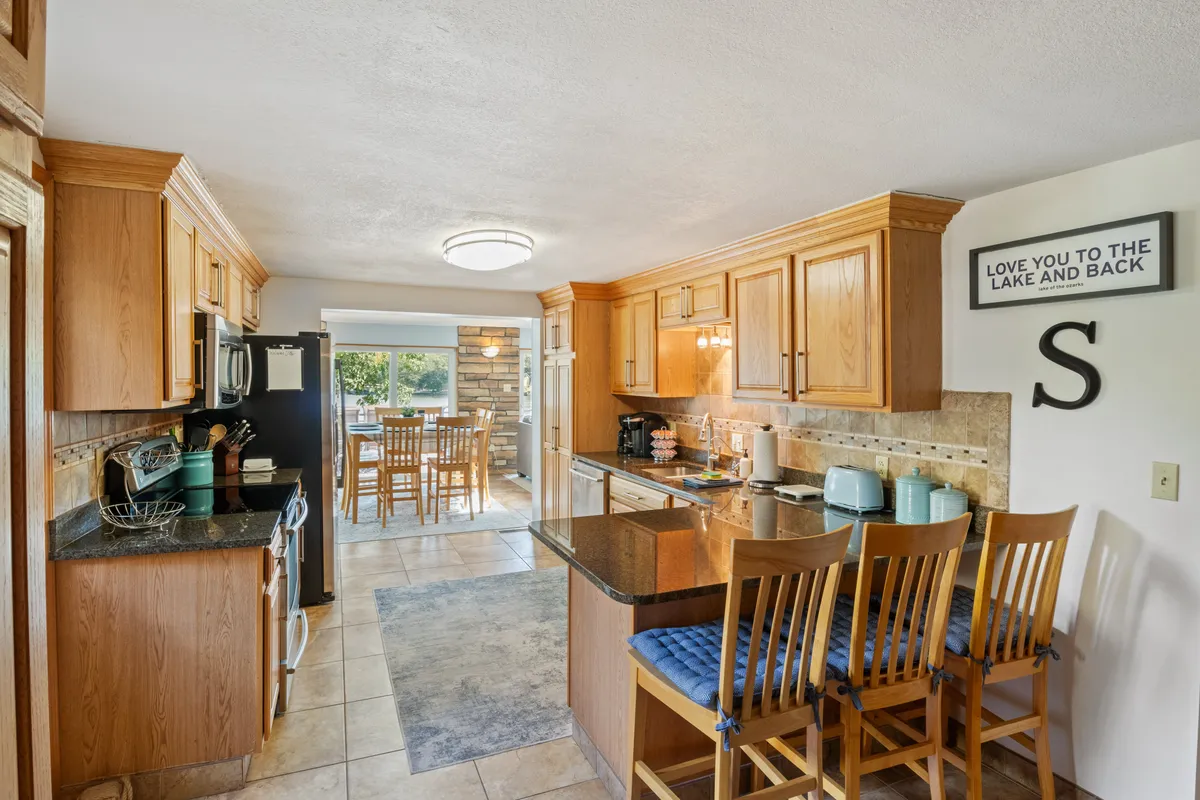 A well-appointed kitchen with a spacious island, stainless steel appliances, rich wood cabinetry, and an adjacent dining area. Large windows bring in ample sunlight and offer glimpses of the surrounding scenery, enhancing the open, communal feel.