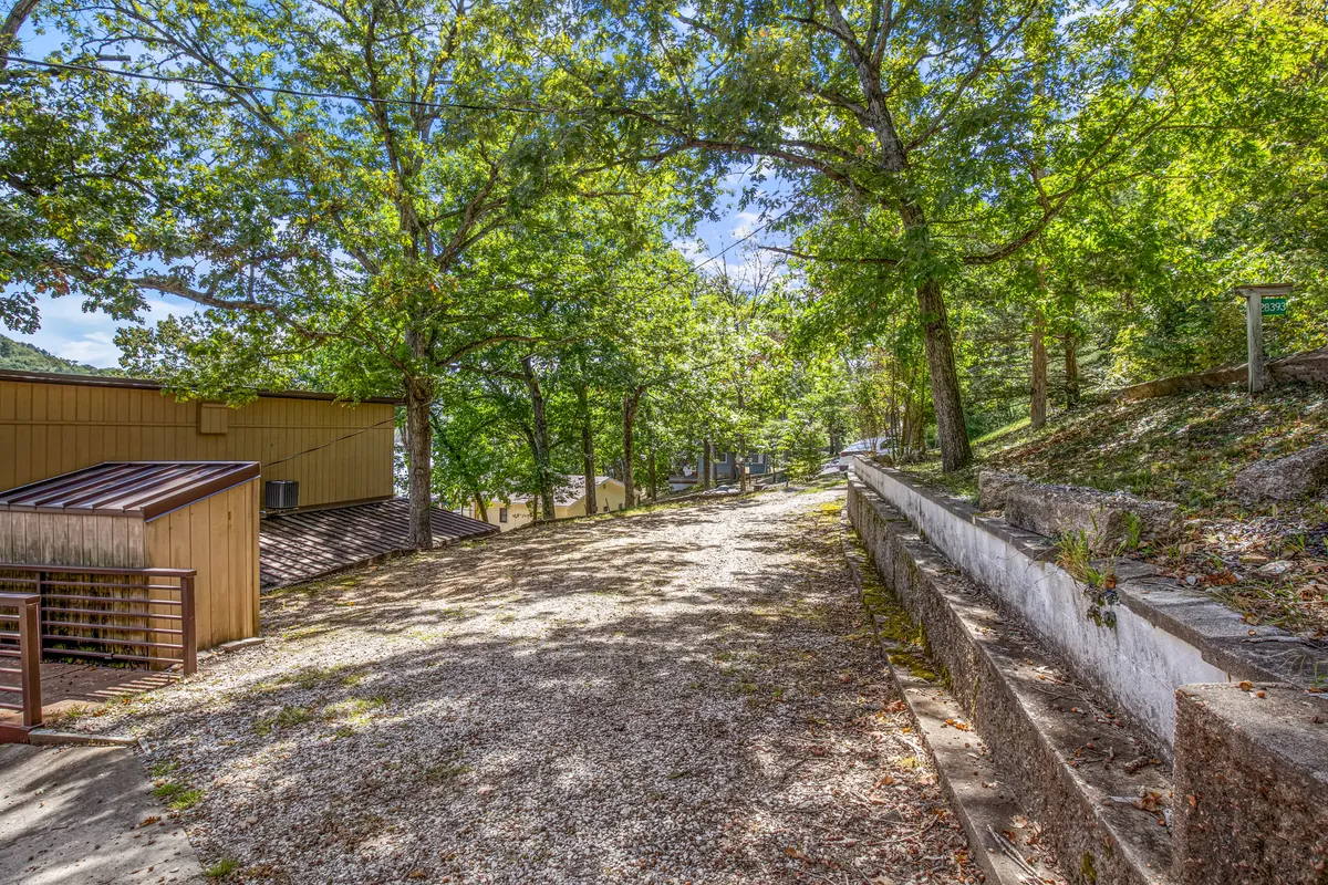 A tranquil exterior scene featuring a gravel path winding under a lush tree canopy, with the building facade visible in the background. The setting invites leisurely strolls and outdoor exploration, evoking the feeling of a peaceful, natural environment suitable for on-leash walks with pets.