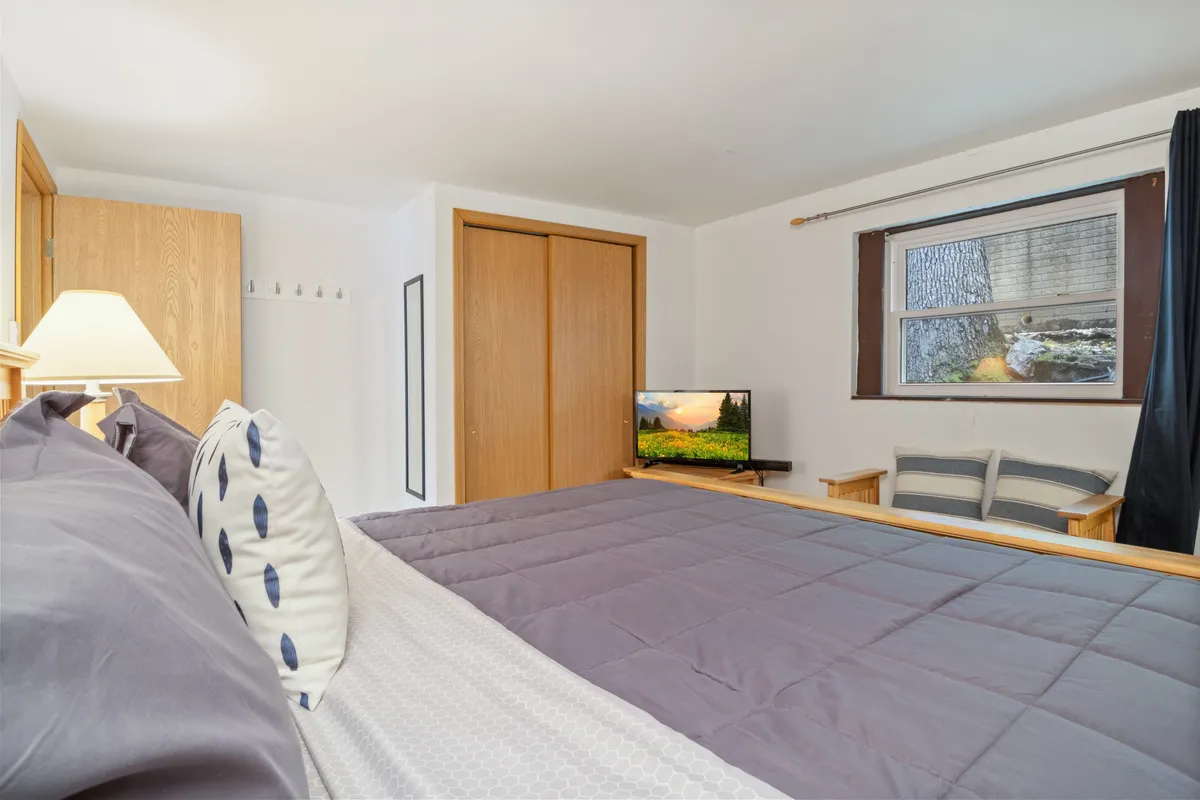 An inviting bedroom featuring gray bedding on a spacious bed, a television, ample natural light through the window, and wooden closet doors. The setting is uncluttered and restful, with touches that suggest both luxury and homey comfort.