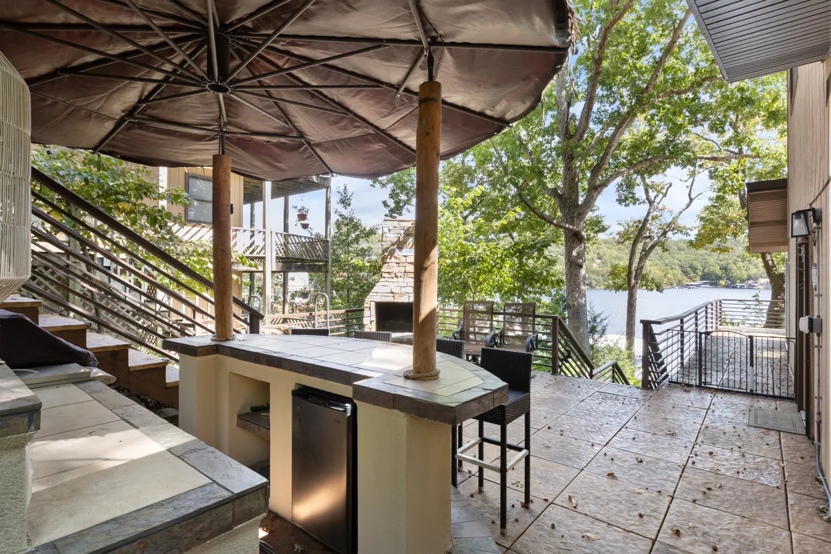 An inviting outdoor bar area with a large umbrella and a direct lake view, featuring wooden stairs and a casual seating arrangement. The scene suggests a vibrant yet relaxed setting for group celebrations, sunset cocktails, or casual gatherings by the water.