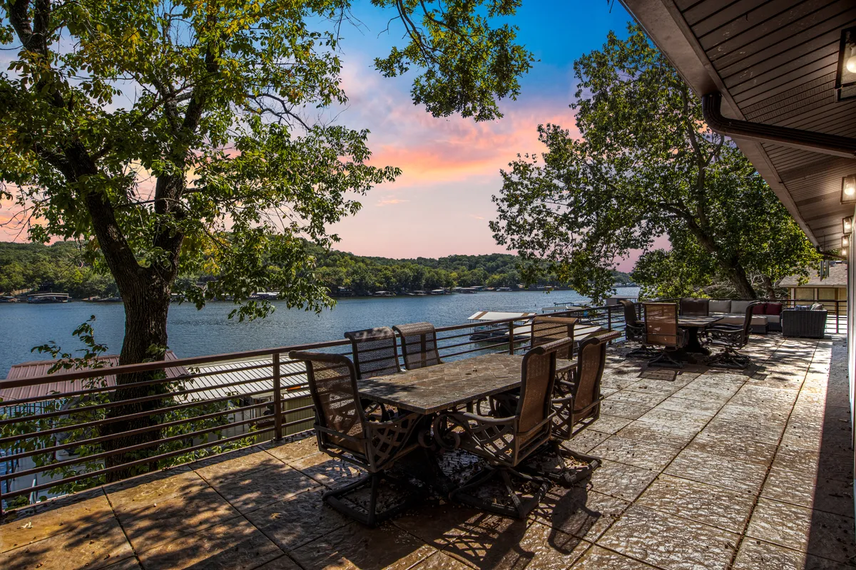 A serene patio scene with a dining table and comfortable chairs set under the open sky, surrounded by lush trees and overlooking a breathtaking lake view. The space is perfect for hosting friends and family or enjoying a quiet sunset moment by the water.
