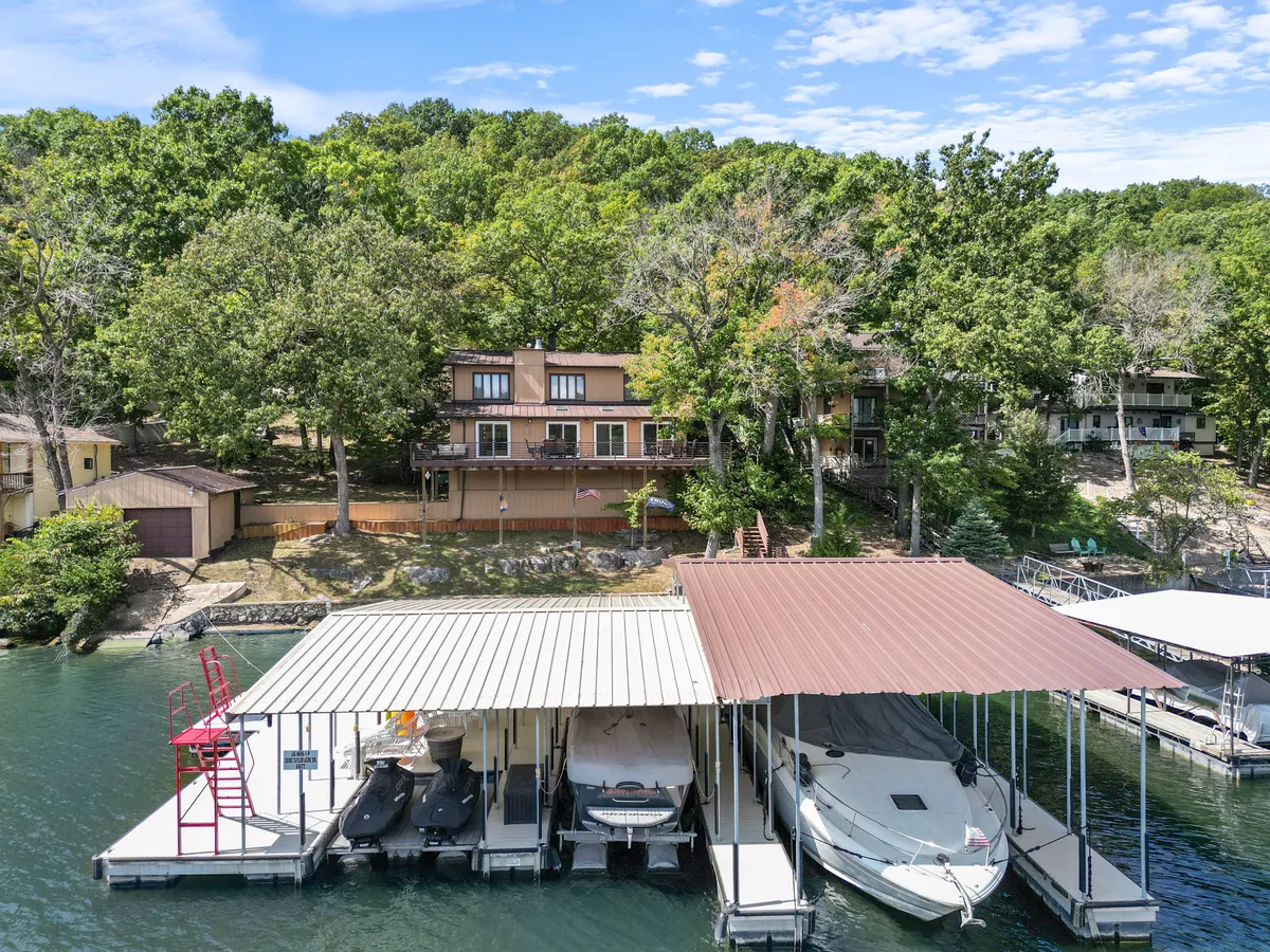A broad view of the lakefront property showing a private dock equipped with boats, surrounded by trees and a balcony visible on the house. The dock sits right on the water’s edge, inviting water activities and relaxation in a serene lakeside setting.