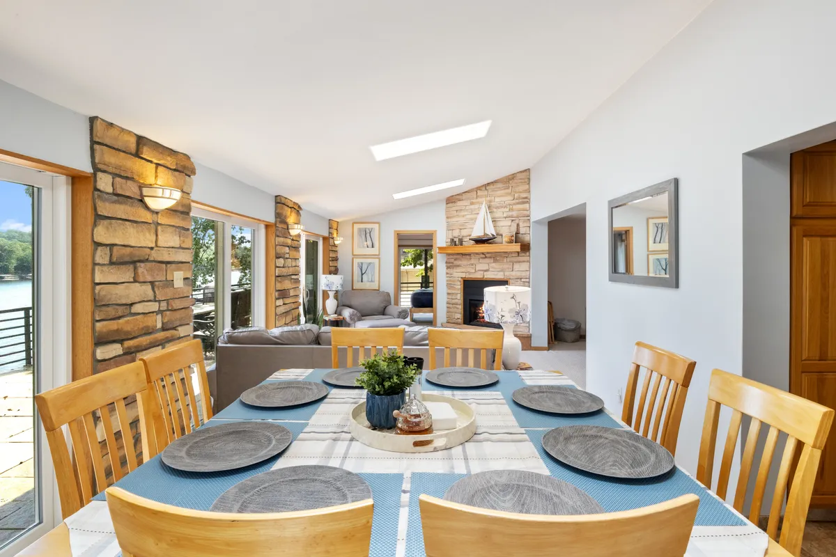 A communal interior space with a central dining table set for family dinners, a striking stone fireplace, large windows framing views of the water, and plush seating nearby. The ambiance is warm, inviting, and ideal for social gatherings.