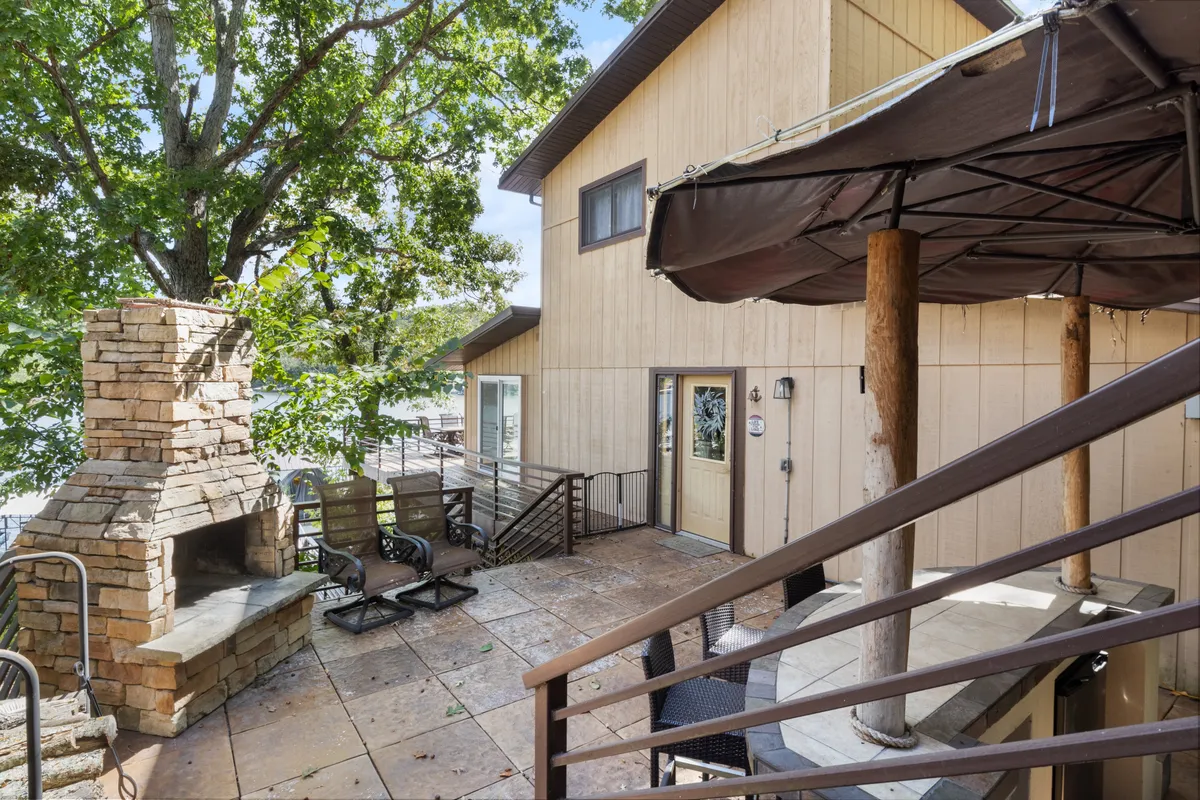 A cozy outdoor seating area arranged around a stone fireplace with wooden railing and glimpses of the house exterior. The setup invites evening gatherings, relaxation, and socializing outdoors, especially as the sun sets.