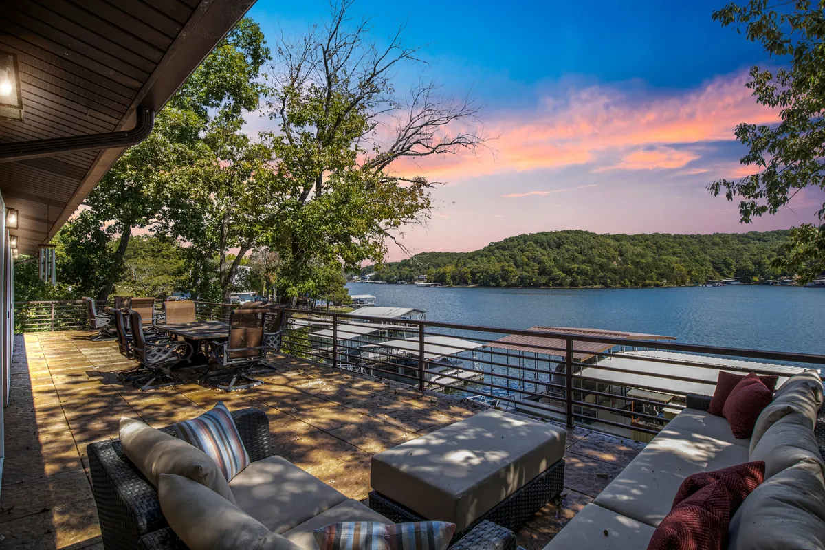 The hero image showcases a stunning lakefront view from the property's expansive outdoor deck. The deck features a comfortable outdoor seating area and a large dining table, all overlooking calm waters with a picturesque sunset. The atmosphere is serene, inviting relaxation, al fresco dining, and social gatherings against a scenic lakeside backdrop.